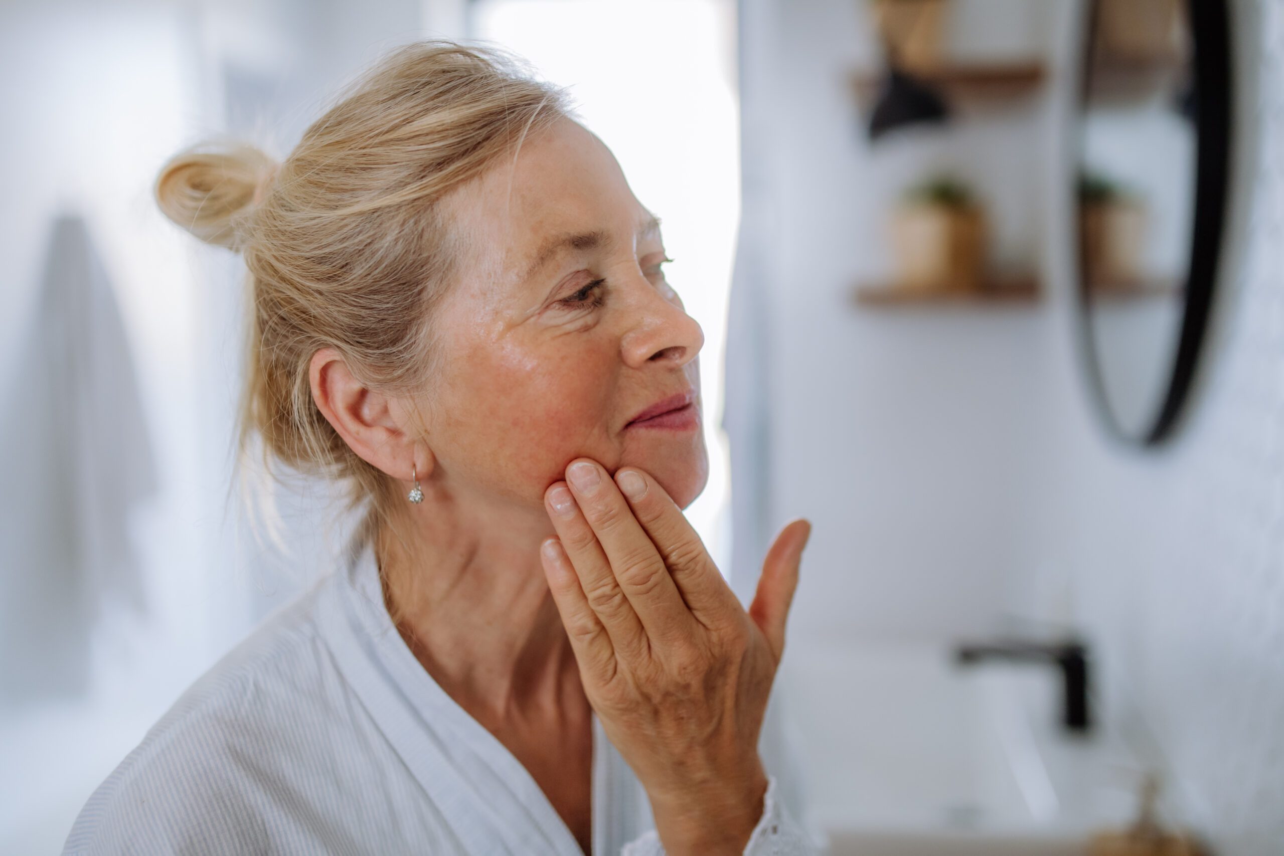 Older woman gently touching her face in the mirror, reflecting on whether rosacea symptoms have changed over time—does rosacea get worse with age?